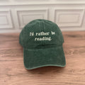 A dark green cotton baseball cap with the phrase 'I'd rather be reading.' embroidered in white on the front, resting on a wooden surface against a white paneled wall.
