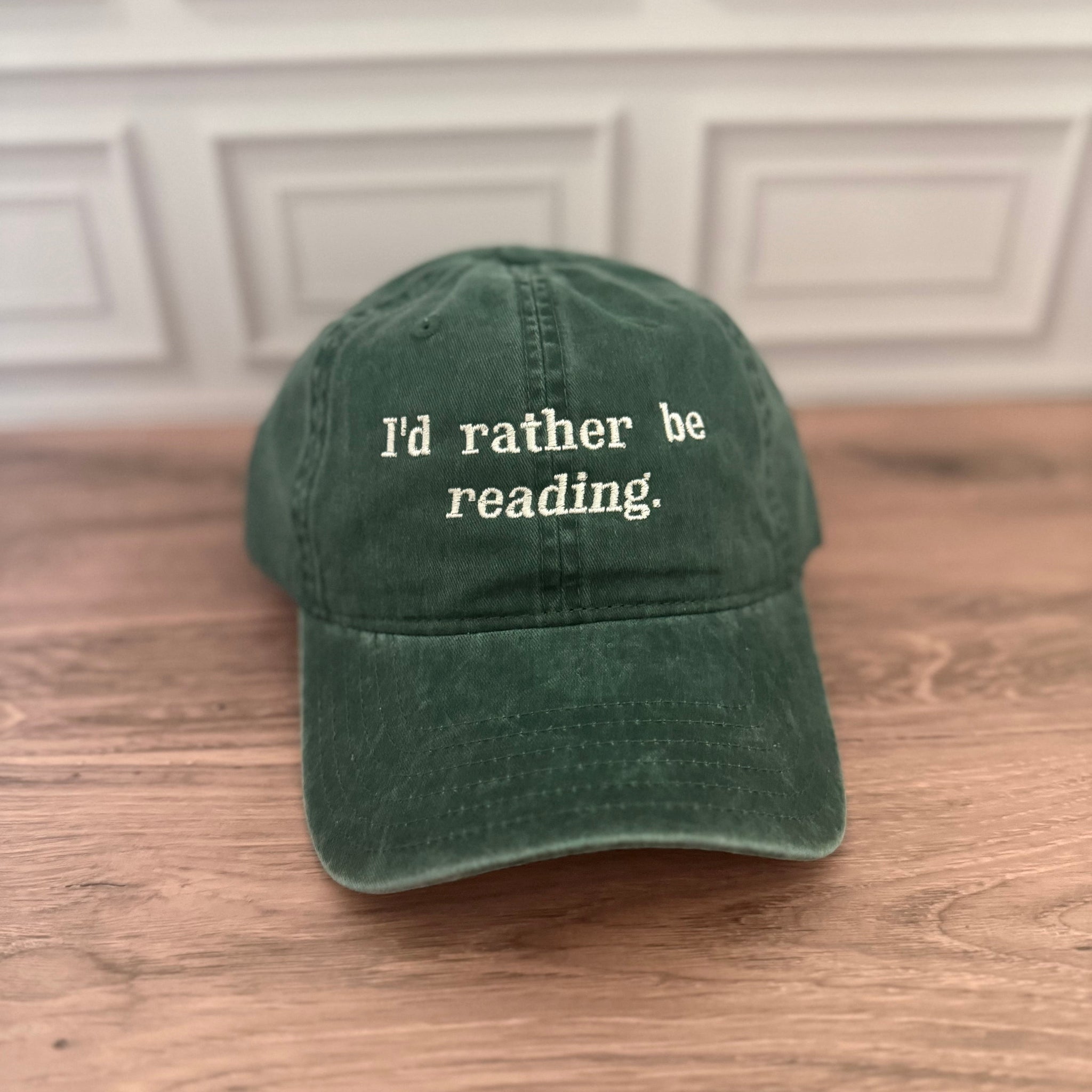 A dark green cotton baseball cap with the phrase 'I'd rather be reading.' embroidered in white on the front, resting on a wooden surface against a white paneled wall.