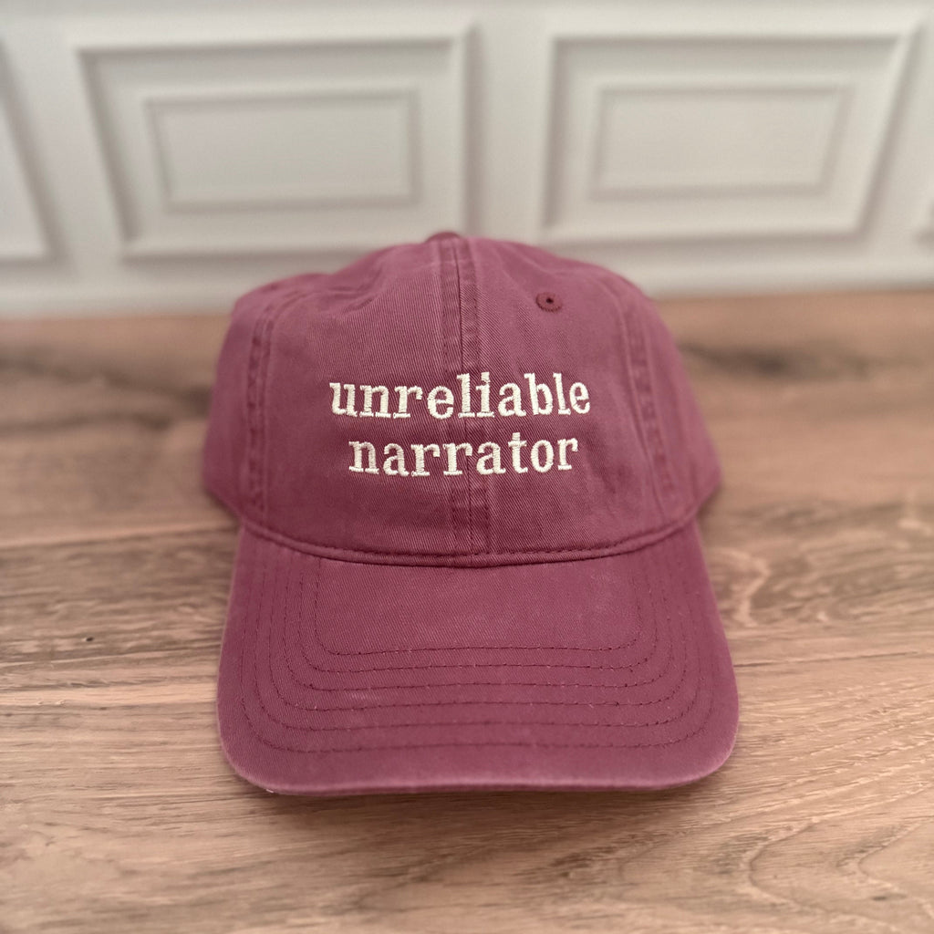 A dusty rose baseball cap with 'unreliable narrator' embroidered in white on the front, resting on a wooden surface with a white paneled wall in the background.